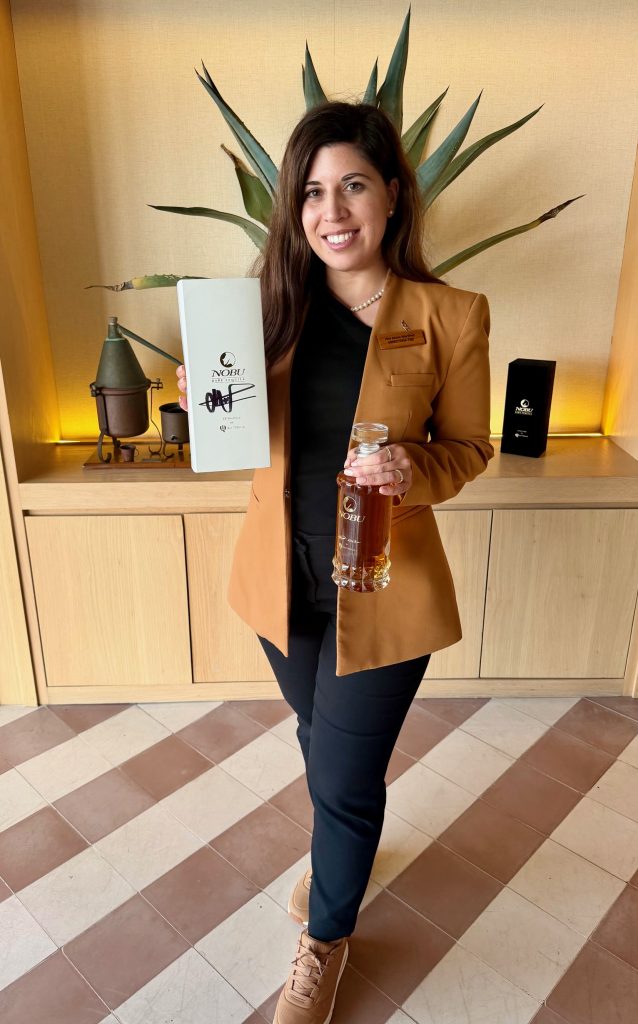 A woman holds a bottle and box of Nobu spirits, standing in front of a decorative plant with a warm, inviting background.