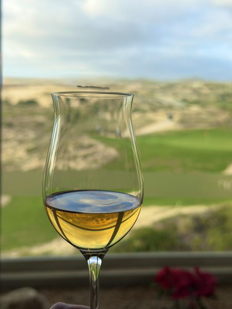 A glass of golden liquid is set against a scenic backdrop of green hills and a cloudy sky at the Nobu Los Cabos masterclass.