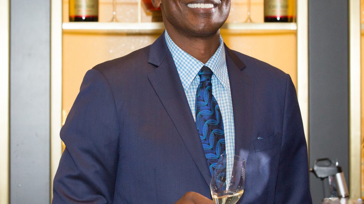 Isiah Thomas smiles while holding a glass of champagne, with wine glasses and champagne bottles displayed in the background.