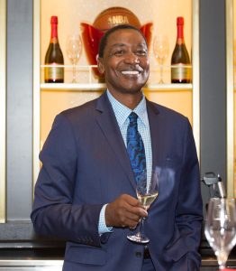 Isiah Thomas smiles while holding a glass of champagne, with wine glasses and champagne bottles displayed in the background.