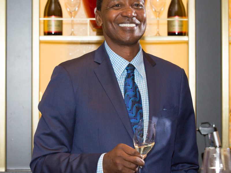 Isiah Thomas smiles while holding a glass of champagne, with wine glasses and champagne bottles displayed in the background.