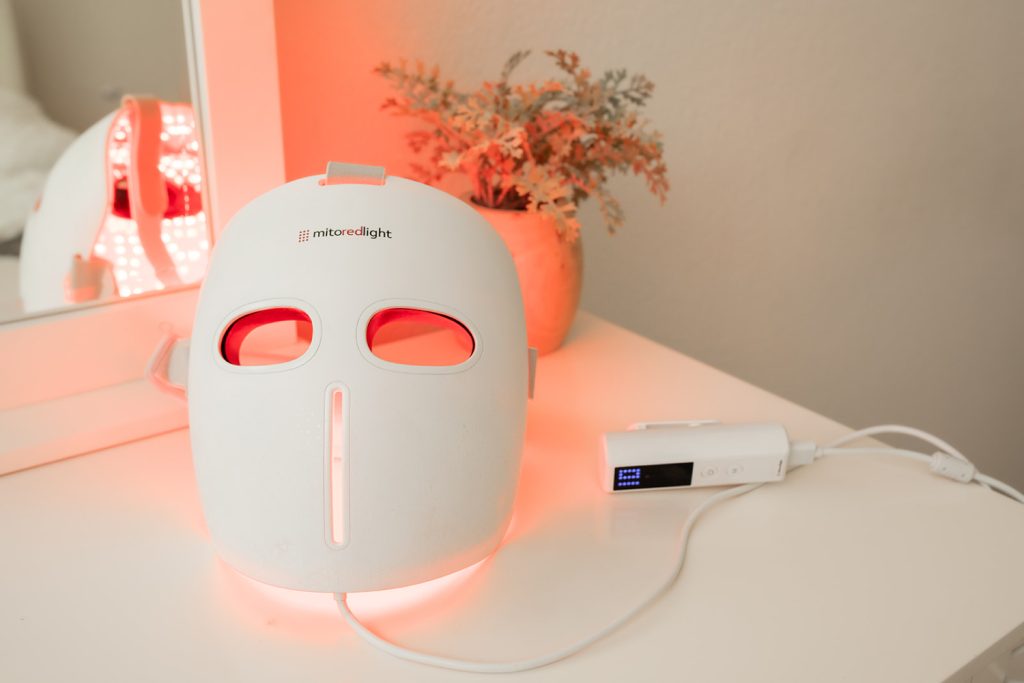 Red light therapy mask on a white surface, with a control device beside it, illuminated by soft red light.