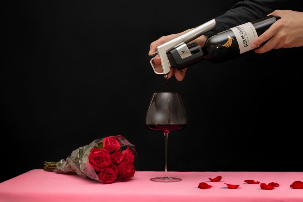 A hand using a Coravin wine system to pour red wine into a glass, with a bouquet of roses and rose petals nearby.