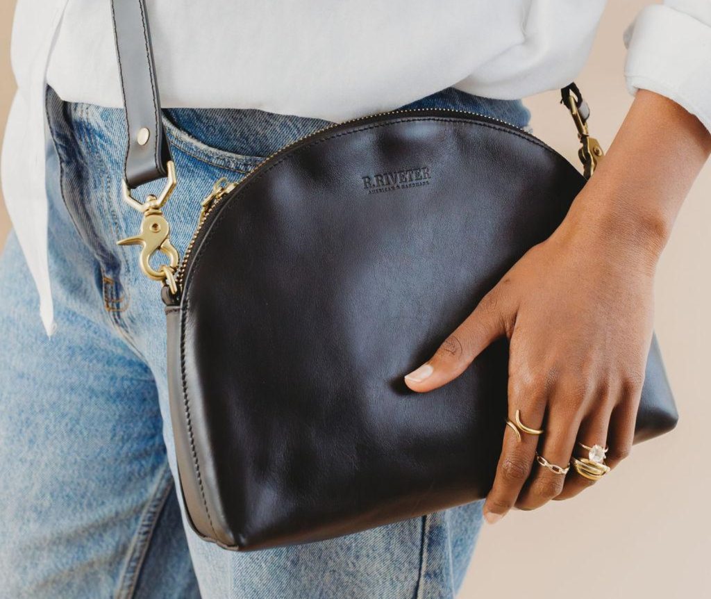 A person holds a sleek black leather bag featuring a gold clasp, paired with denim and stylish rings on their fingers.