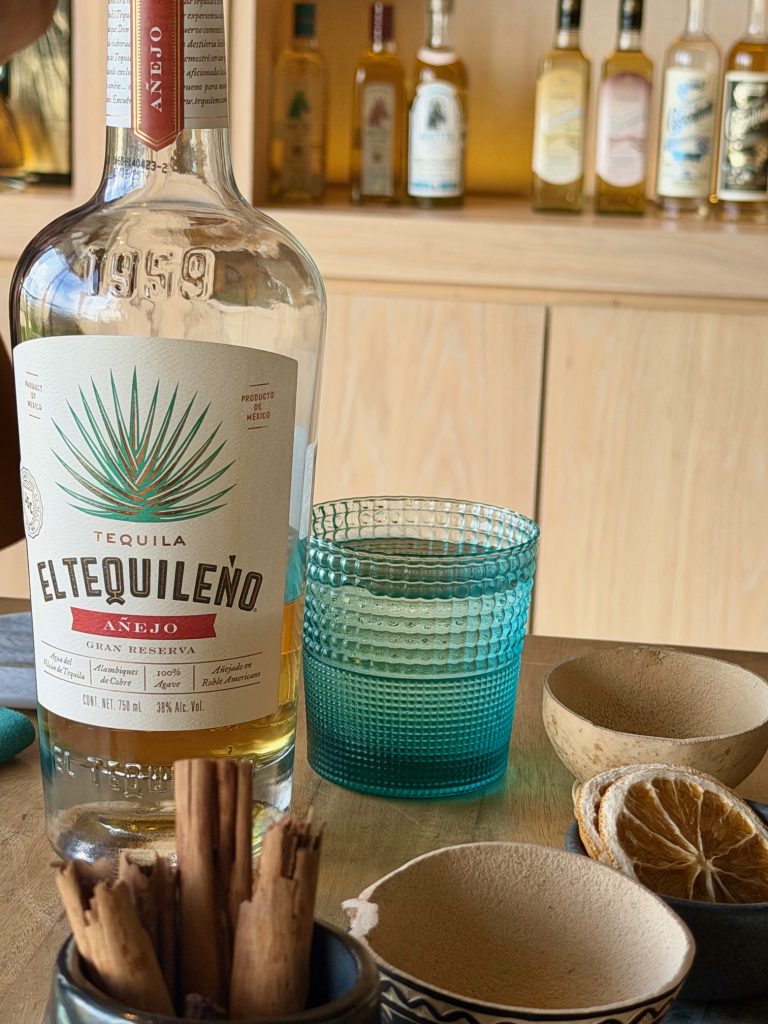 Tequila El Tequileno Añejo with a turquoise glass, cinnamon sticks, and dried orange slices on a wooden table.