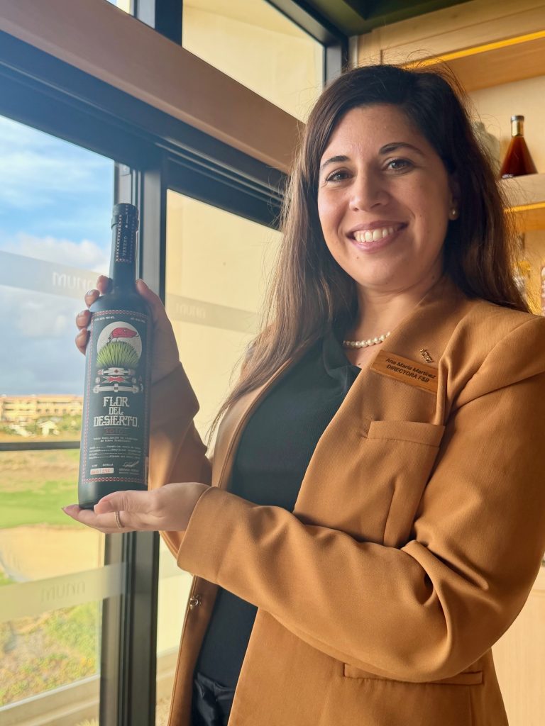 A woman in a brown blazer holds a bottle labeled "Flor del Desierto" at the Beyond The Agave Masterclass at Nobu Los Cabos.