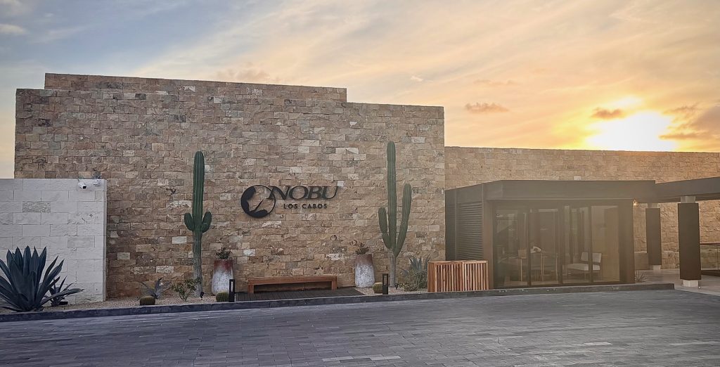 The entrance of Nobu Los Cabos featuring natural stone walls, cacti, and a sunset backdrop.