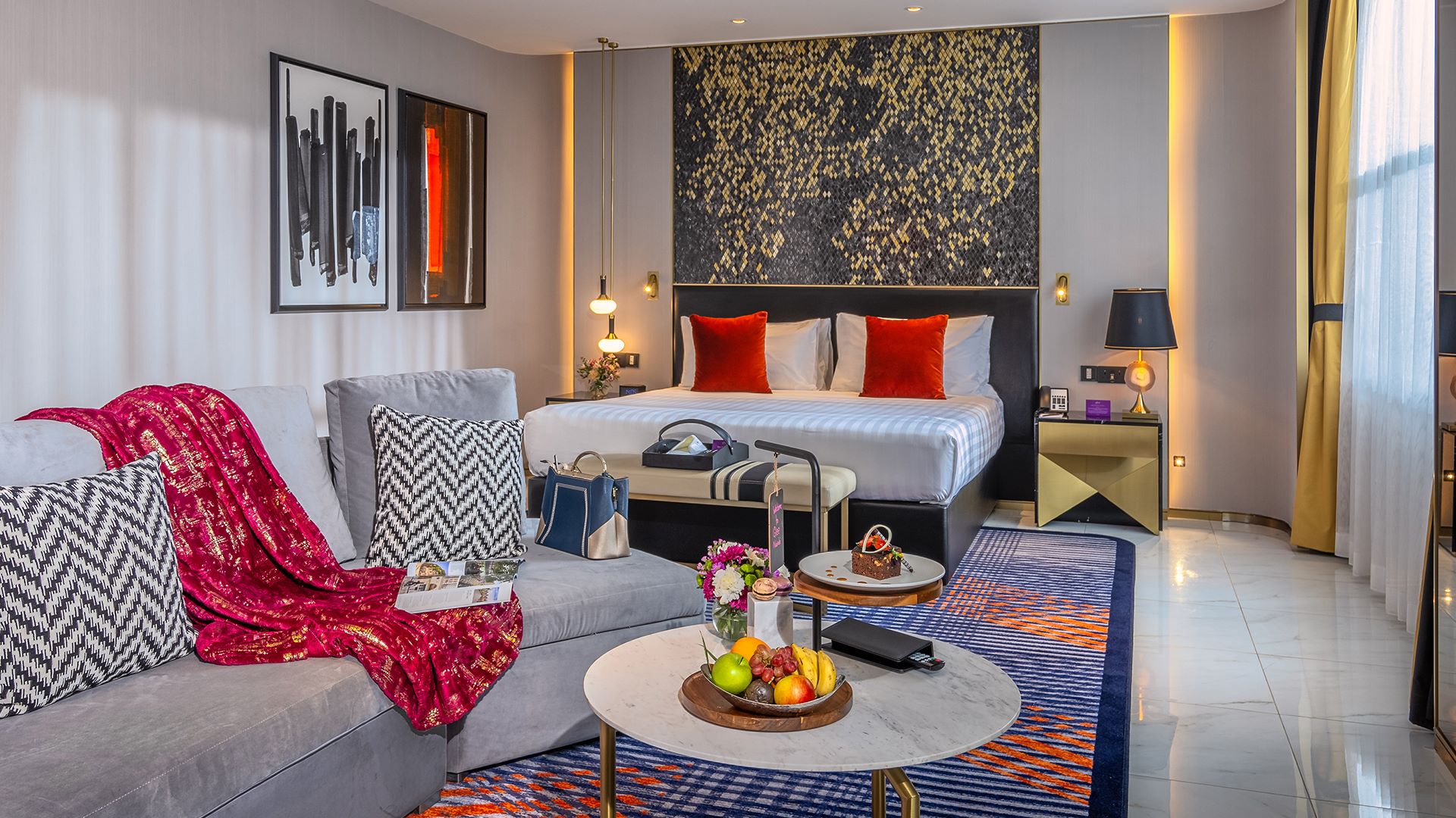 Luxurious hotel room with a queen bed, stylish decor, seating area, and a table with fresh fruit and pastries.