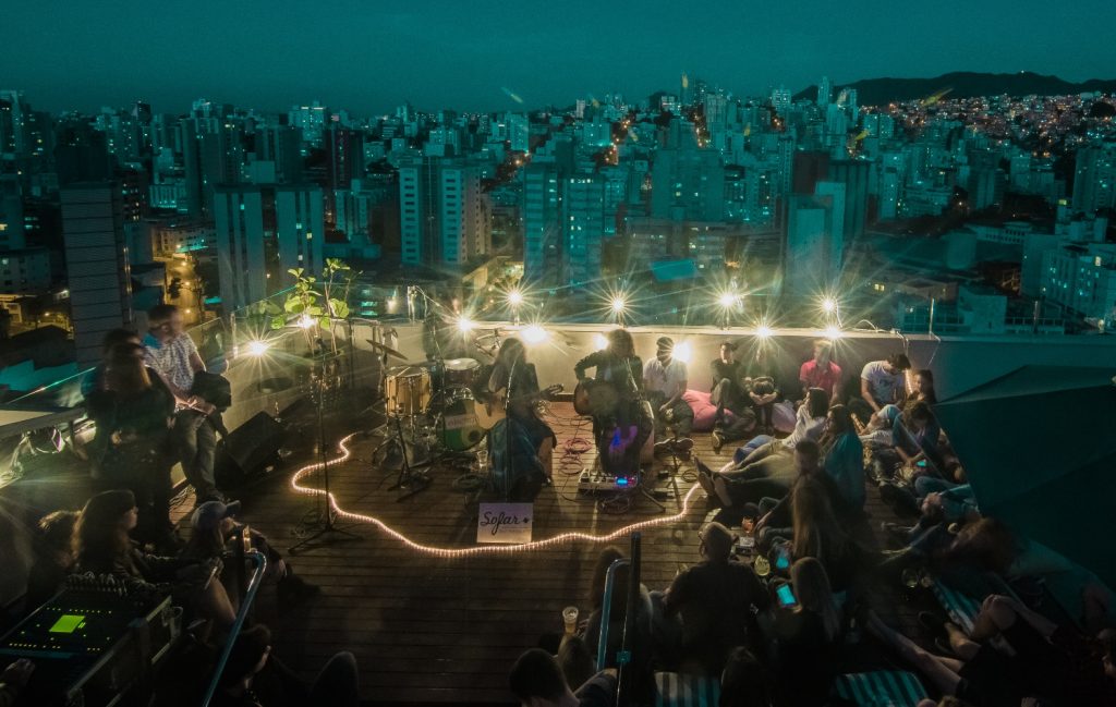 Rooftop concert scene with performers, audience enjoying the view of the city illuminated at night.