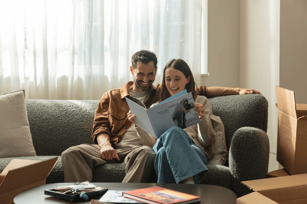 A couple happily reading a photo book together while seated on a cozy sofa, surrounded by unpacked boxes.