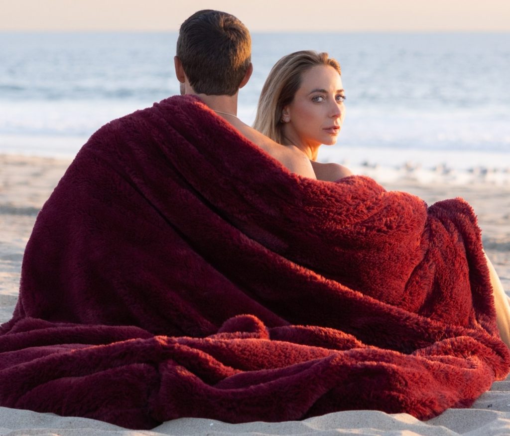 Couple wrapped in a cozy maroon blanket on the beach, gazing at the ocean during sunset.
