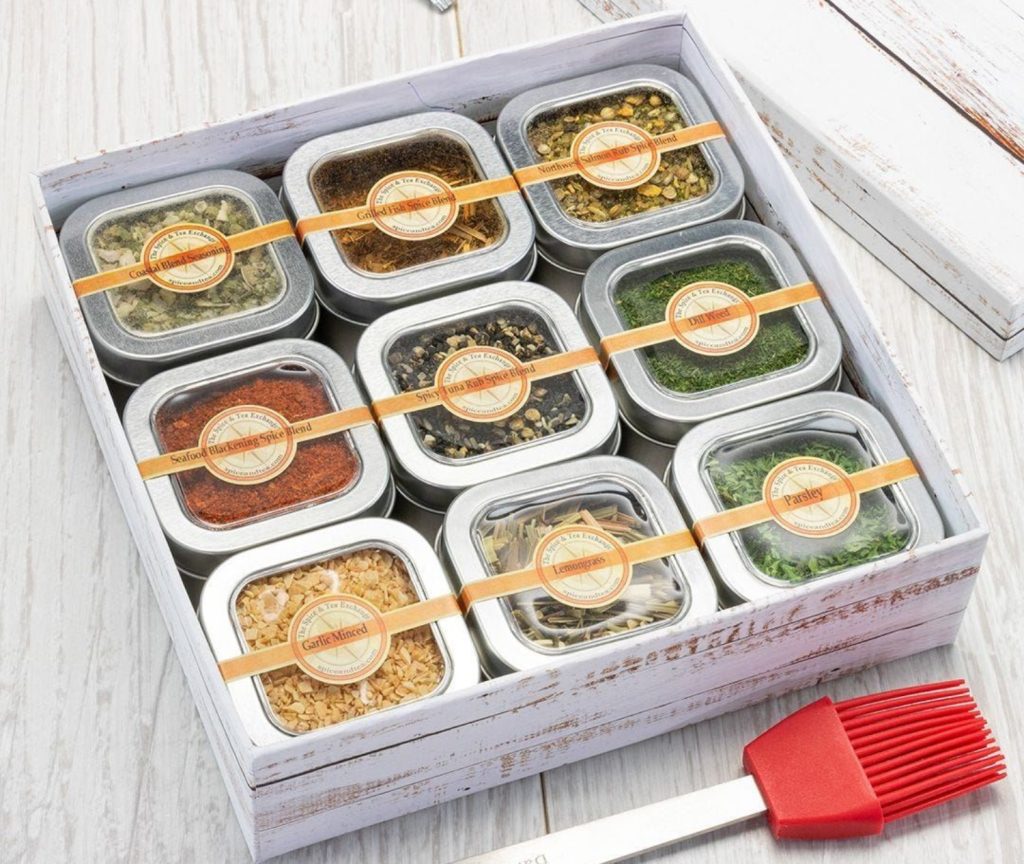 Nine spice blends in a decorative tin gift box, including seafood blackening, dill, and garlic, arranged on a wooden surface.