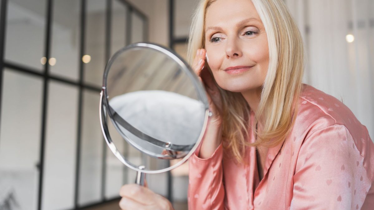 Woman in a silk top applying skincare while looking in a mirror, reflecting self-care and beauty routines.