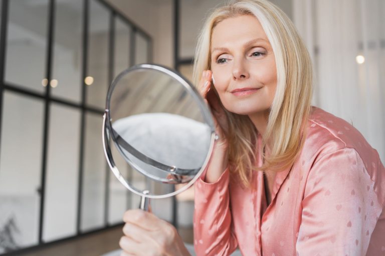 Woman in a silk top applying skincare while looking in a mirror, reflecting self-care and beauty routines.
