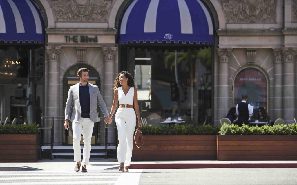 A couple walks hand in hand in front of a restaurant with a blue-and-white awning, enjoying a sunny day in LA.