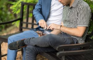 Two men discuss content on a tablet while seated on a bench, surrounded by greenery.