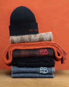 A navy knitted beanie sits atop colorful stacked scarves in various textures against a vibrant orange background.