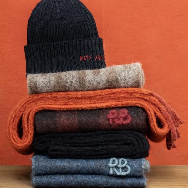 A navy knitted beanie sits atop colorful stacked scarves in various textures against a vibrant orange background.