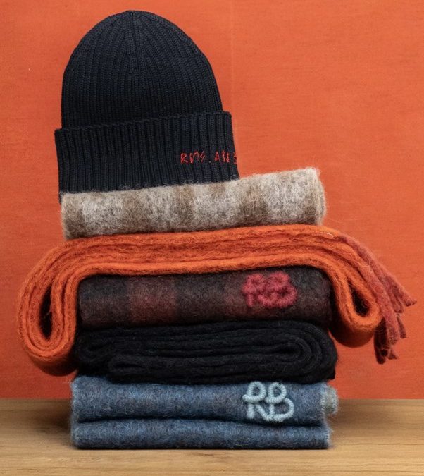 A navy knitted beanie sits atop colorful stacked scarves in various textures against a vibrant orange background.