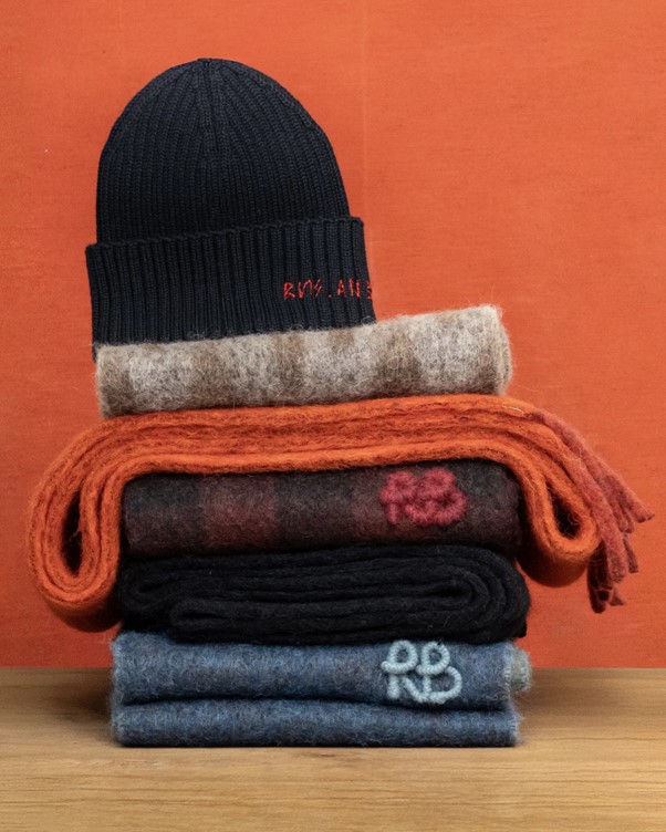 A navy knitted beanie sits atop colorful stacked scarves in various textures against a vibrant orange background.