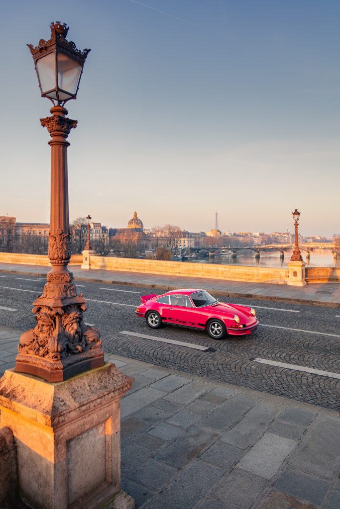 A vibrant pink Porsche 911 Carrera RS 2.7 driving along a scenic cobblestone street with historic buildings and lampposts.