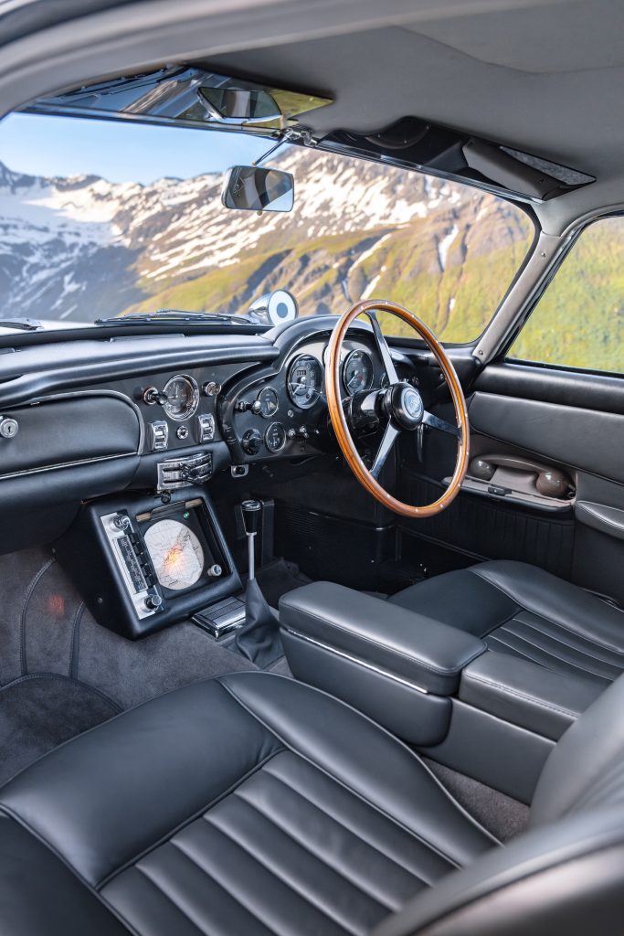 Interior view of an Aston Martin DB5 showcasing black leather seats and a classic dashboard against a scenic background.