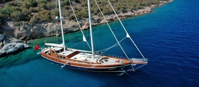 A wooden gulet moored in turquoise waters near a rocky coastline in Turkiye, showcasing its sails and Turkish flag.