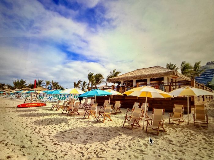 Beach chairs and umbrellas set up on sandy shore with a beach bar and palm trees under a partly cloudy sky.