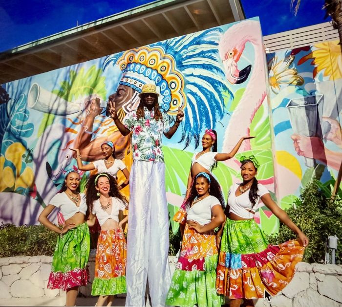 A stilt walker poses with six women in colorful skirts in front of a vibrant mural at Royal Caribbean's Royal Beach Club.