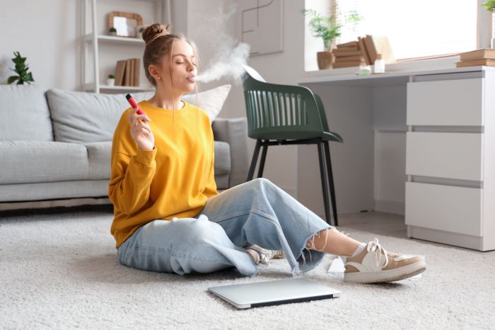 Woman sitting on the floor, vaping with a device, in a cozy room with a laptop and plants in the background.