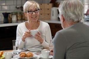 An elderly woman with glasses enjoys coffee while chatting with a man at a breakfast table filled with pastries.