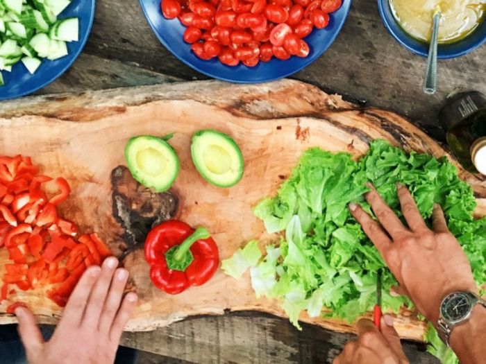 Fresh vegetables being prepared on a wooden cutting board, with hands chopping red bell peppers and lettuce.