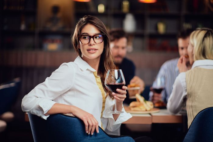 A woman in stylish eyeglasses enjoys a glass of red wine while socializing with friends at a trendy restaurant.