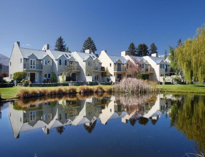 Reflective lake view of resort buildings surrounded by trees and greenery under a clear blue sky.