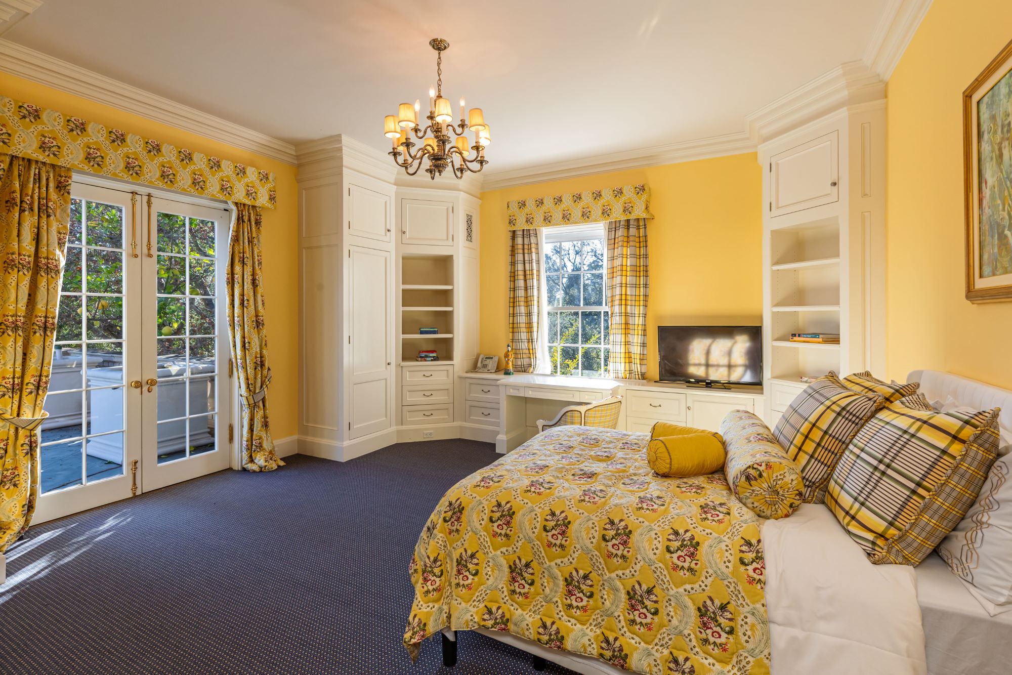 Brightly decorated bedroom featuring yellow walls, floral bedding, a chandelier, and a view through French doors.