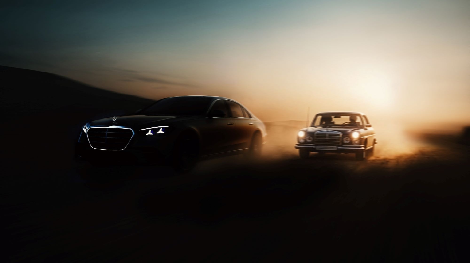 A modern Mercedes-Benz sedan and a classic model drive side by side in a dusty landscape at sunset.