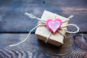 Wrapped package adorned with a pink heart on top, tied with string, against a wooden background.