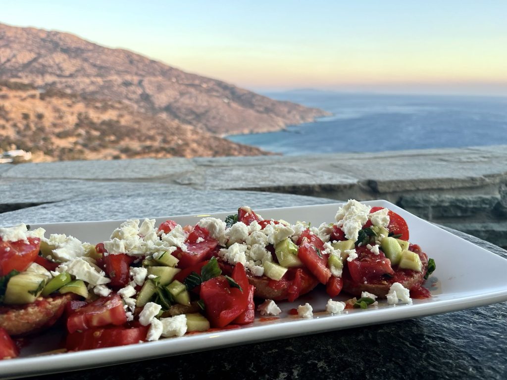 Fresh Greek salad with tomatoes, cucumbers, and feta cheese, served against a stunning coastal sunset view.