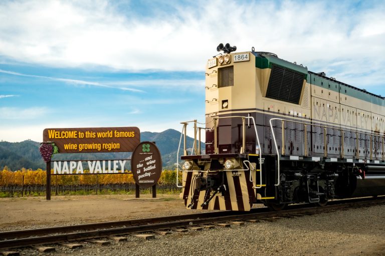 Napa Valley Wine Train locomotive stands near a welcome sign for the Napa Valley wine growing region amidst vineyards.