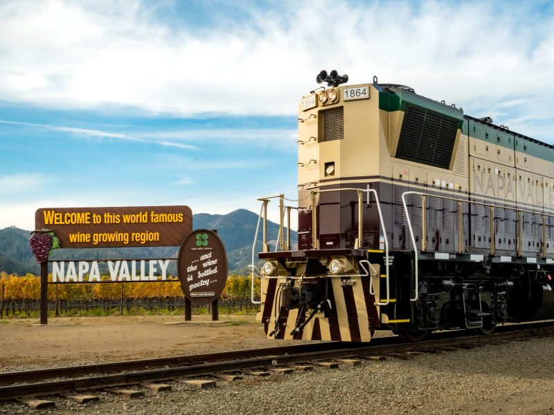 Napa Valley Wine Train locomotive stands near a welcome sign for the Napa Valley wine growing region amidst vineyards.
