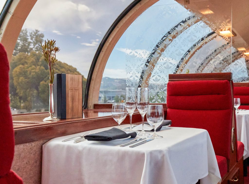 Elegant dining setup inside the Napa Valley Wine Train, featuring red chairs, glassware, and a scenic view through large windows.