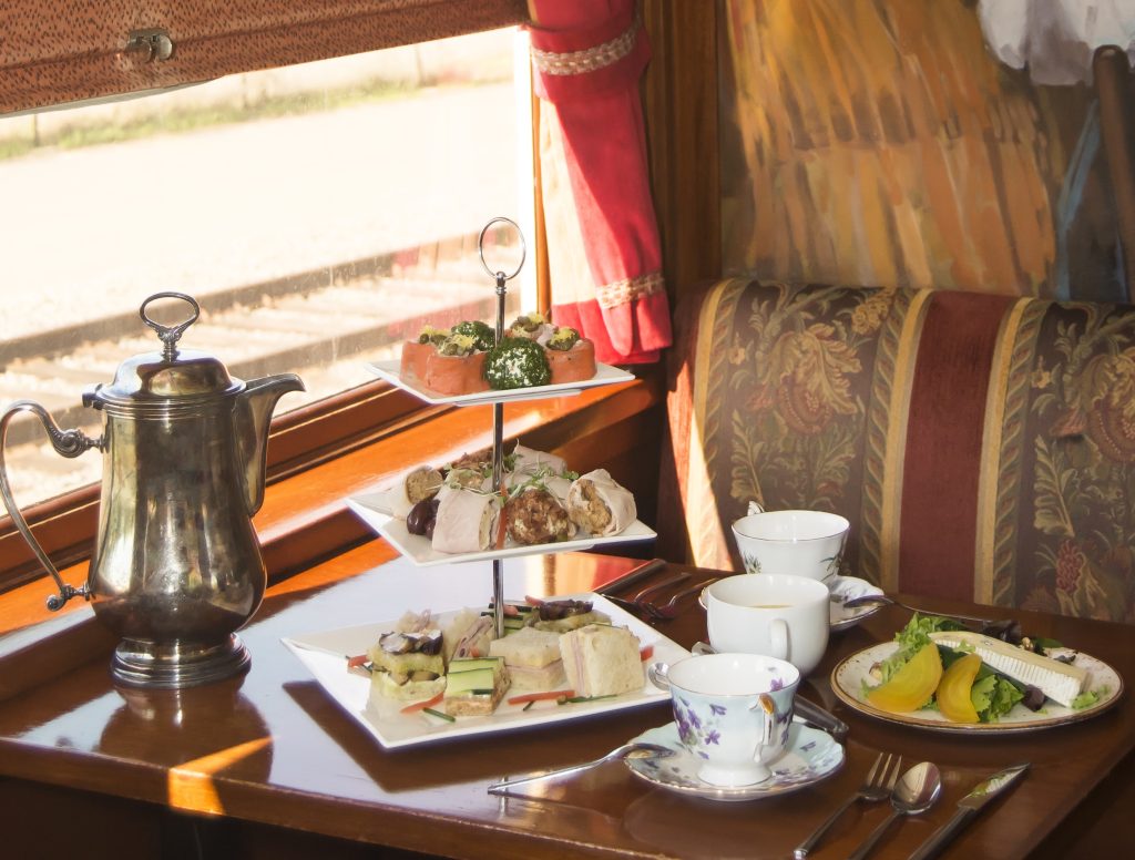 Three-tiered afternoon tea display with silver teapot, assorted sandwiches, pastries, and garnishes on a dining table in a train car.