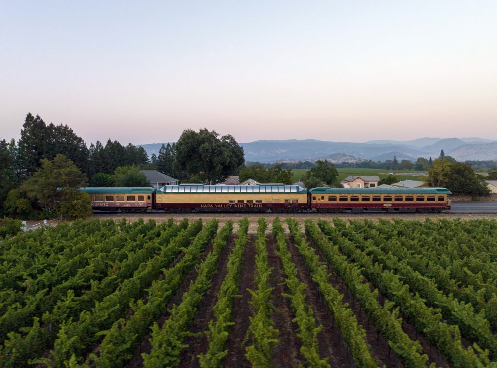 Napa Valley Wine Train passes through lush vineyards with a scenic backdrop of mountains at dusk.