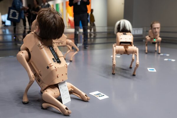 Robotic sculptures with human-like heads are arranged on a gallery floor, exploring themes of technology and consumerism.
