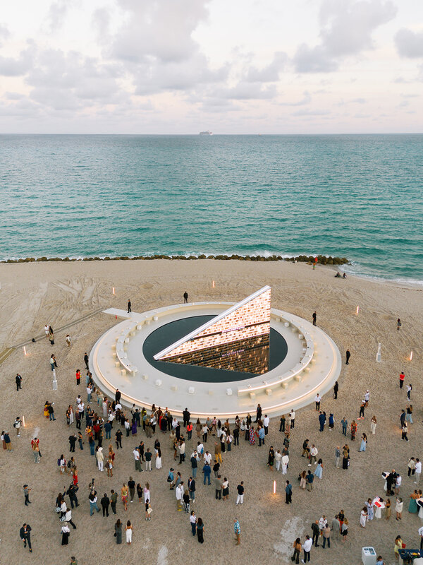 Crowd gathered around a contemporary sculpture on a beach, with the ocean and cloudy sky in the background at Art Basel Miami Beach 2025.