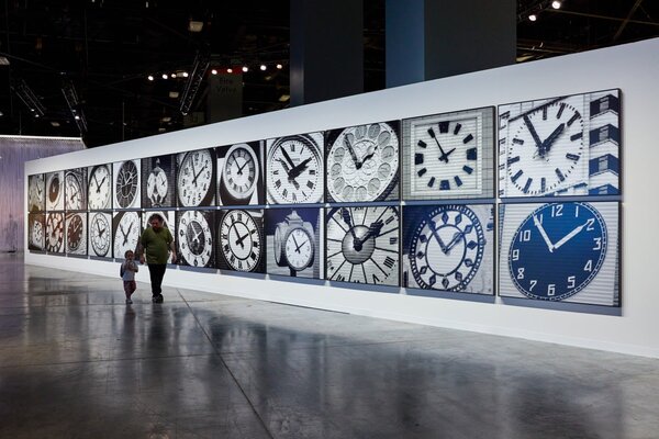 A woman and child walk past a large wall installation featuring various clock designs at Art Basel Miami Beach 2025.