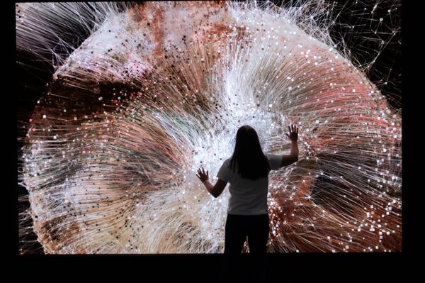 A person stands in front of a large, colorful digital artwork with dynamic lines and dots, engaging with the immersive experience.