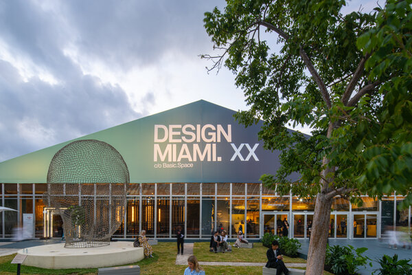 Design Miami venue with a large sign, featuring visitors and a sculpture in the foreground, against a partly cloudy sky.