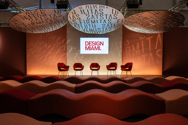 Design Miami stage featuring four red chairs in front of a screen displaying "DESIGN MIAMI." with abstract patterned overhead lights.