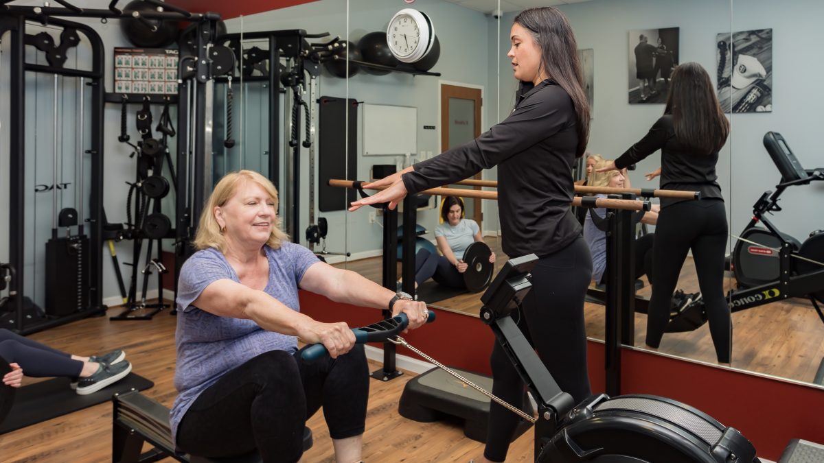 A fitness trainer guides a woman on a rowing machine, while others follow a workout in the background of a gym.